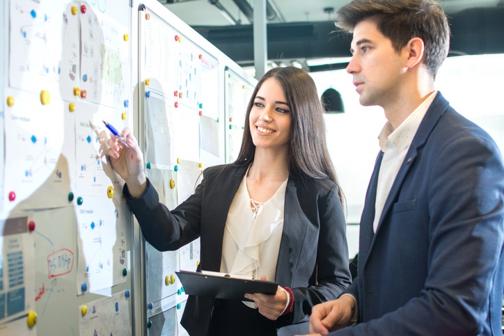 A man and a woman, both in business suits, standing looking at a whiteboard. The woman is teaching the man something