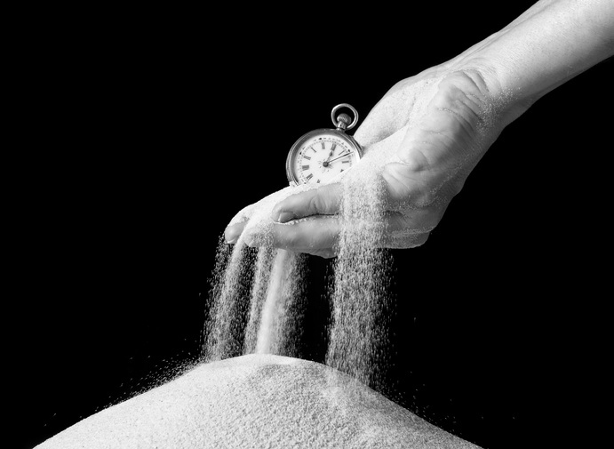 A hand holding a small clock with sand slipping through the fingers into a pile of sand below