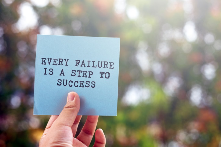 A person's hand holding a sticky note that says "Every failure is a step to success", set against an outdoor background