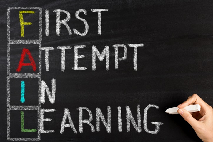 The words "first attempt in learning" written on a chalkboard in white chalk with the first letter of each word written in color and a box drawn around the letter. These letters spell out the word "fail"