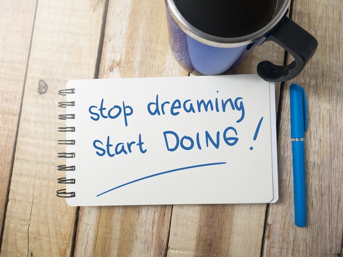 A cup of coffee, a notebook, and a pen on a table. "Stop dreaming. Start doing!" is written in the notebook