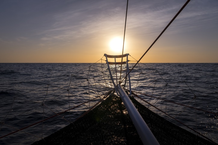 A sailboat on the ocean heading towards the sun on the horizon