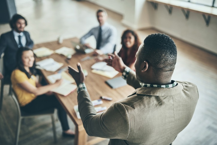 A person standing leading a meeting with 4 people sitting at a desk taking notes and looking engaged