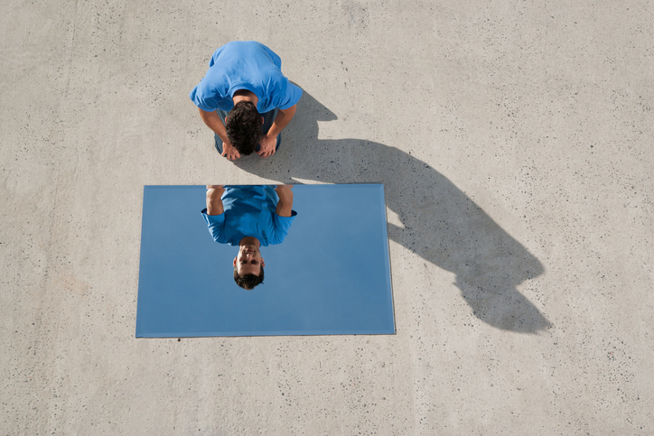 A mirror on the ground with a man crouching down looking into it at his reflection