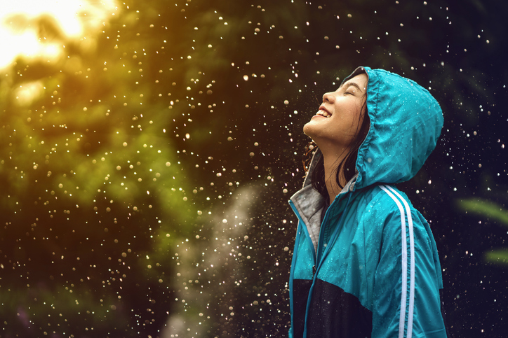 Photo of a young woman in a raincoat standing outside in a light rain while the sun shines through. Her head is tilted up to the sky and she is smiling