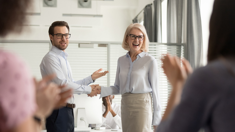A man and a woman standing at the front of a room shaking hands. The man is pointing to the woman in acknowledgement and the rest of the people in the room are applauding